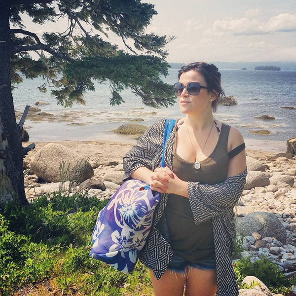 woman wearing sunglasses holding beach bag standing in front of ocean stares off into distance